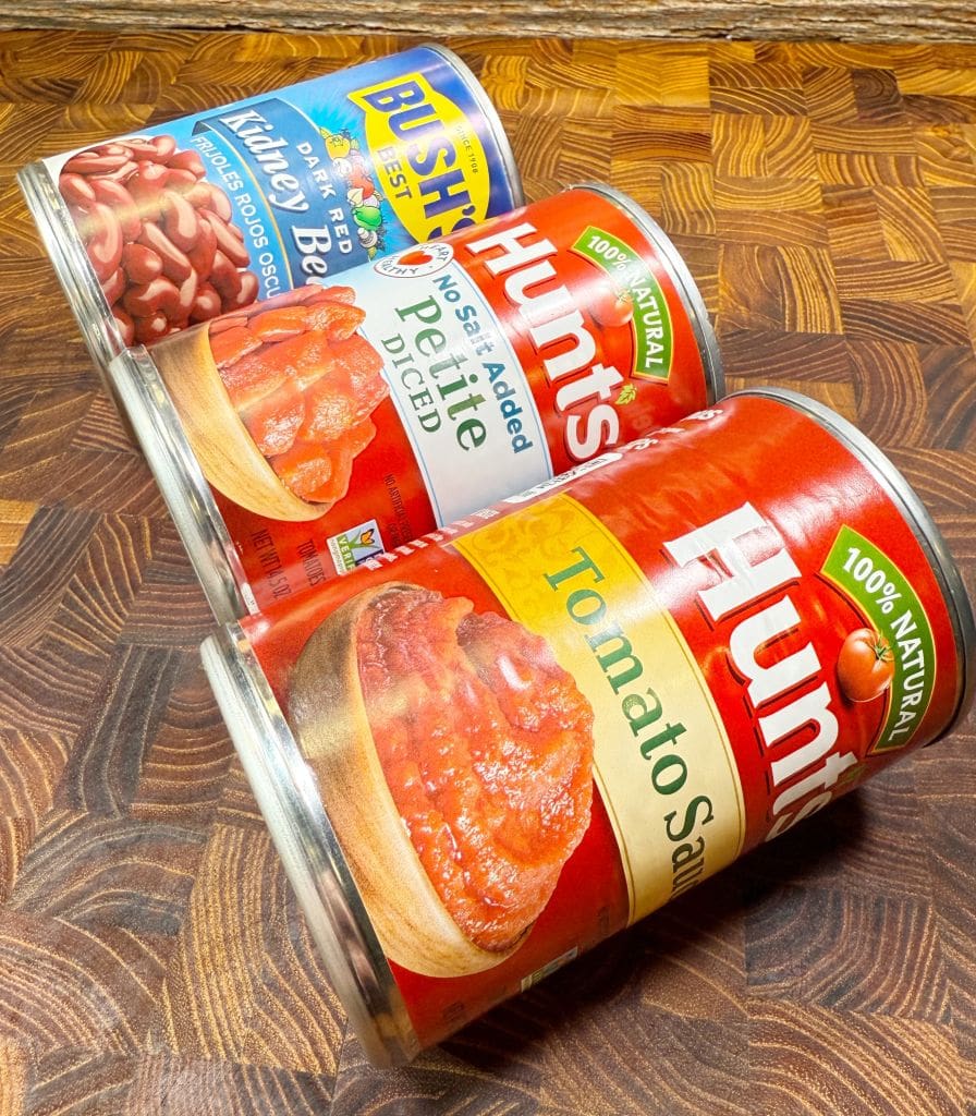 Three cans on a wooden surface: one of Bushs Best kidney beans, one of Hunts no-salt-added petite diced tomatoes, and one of Hunts tomato sauce, all labeled as 100% natural.