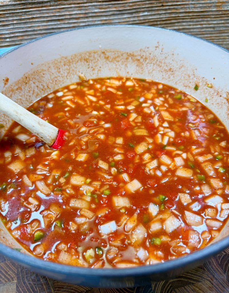 A pot filled with a rich, chunky red sauce containing diced vegetables and spices for the sloppy joe recipe. A wooden spoon rests inside the pot, slightly stained by the sauce. The pot sits on a patterned wooden surface.