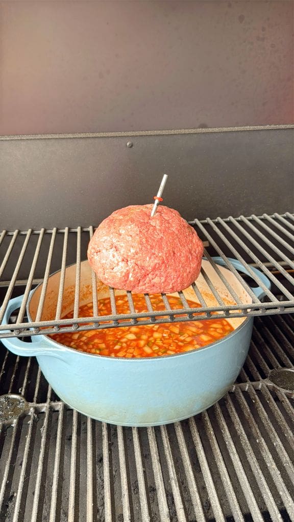 A large raw meatloaf is shaped into a dome and placed on an upper grill rack, with a temperature probe inserted. Below, a blue pot filled with baked beans simmers on a barbecue grill.