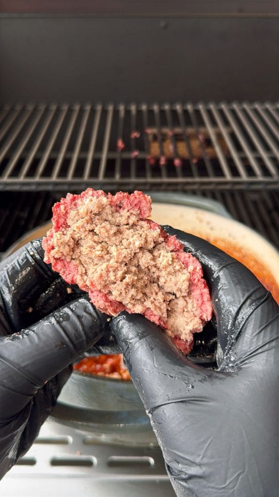 Hands wearing black gloves hold a partially cooked hamburger patty over a grill. The patty shows a mix of raw red and cooked brown meat. A pan is visible on the grill in the background.