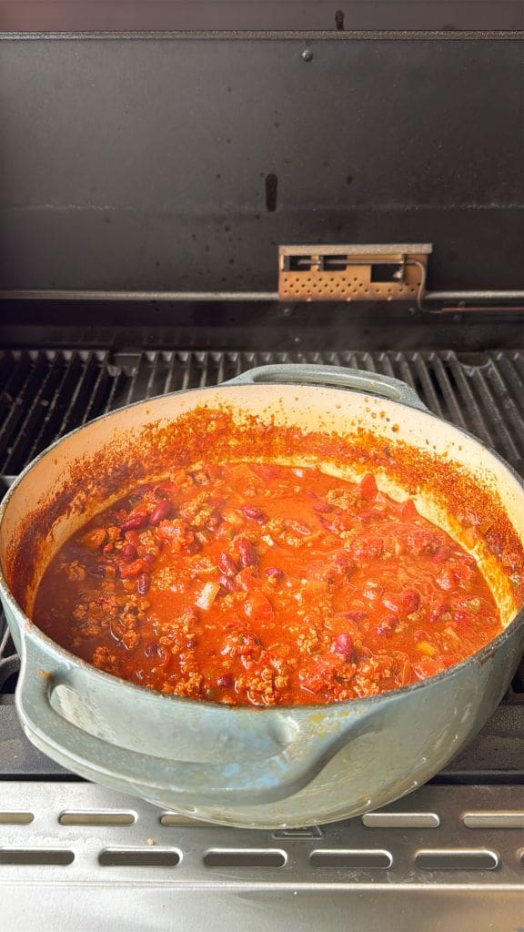 A pot of chili cooking on a stovetop, filled with red kidney beans, tomatoes, and ground meat in a rich red sauce. The pot is set on a metal grill surface.