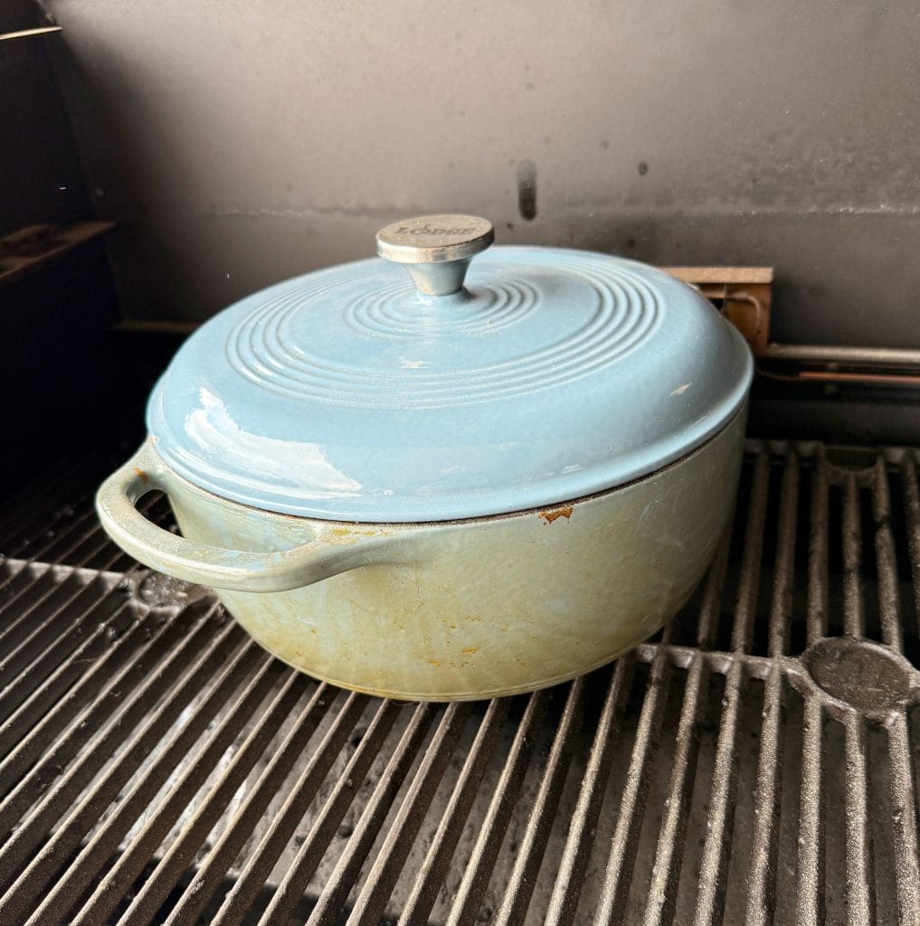 A light blue cast iron pot with a lid sits on a black grill grate. The pot has a round handle on the lid and a handle on the side. The surface of the pot shows signs of use with some discoloration.