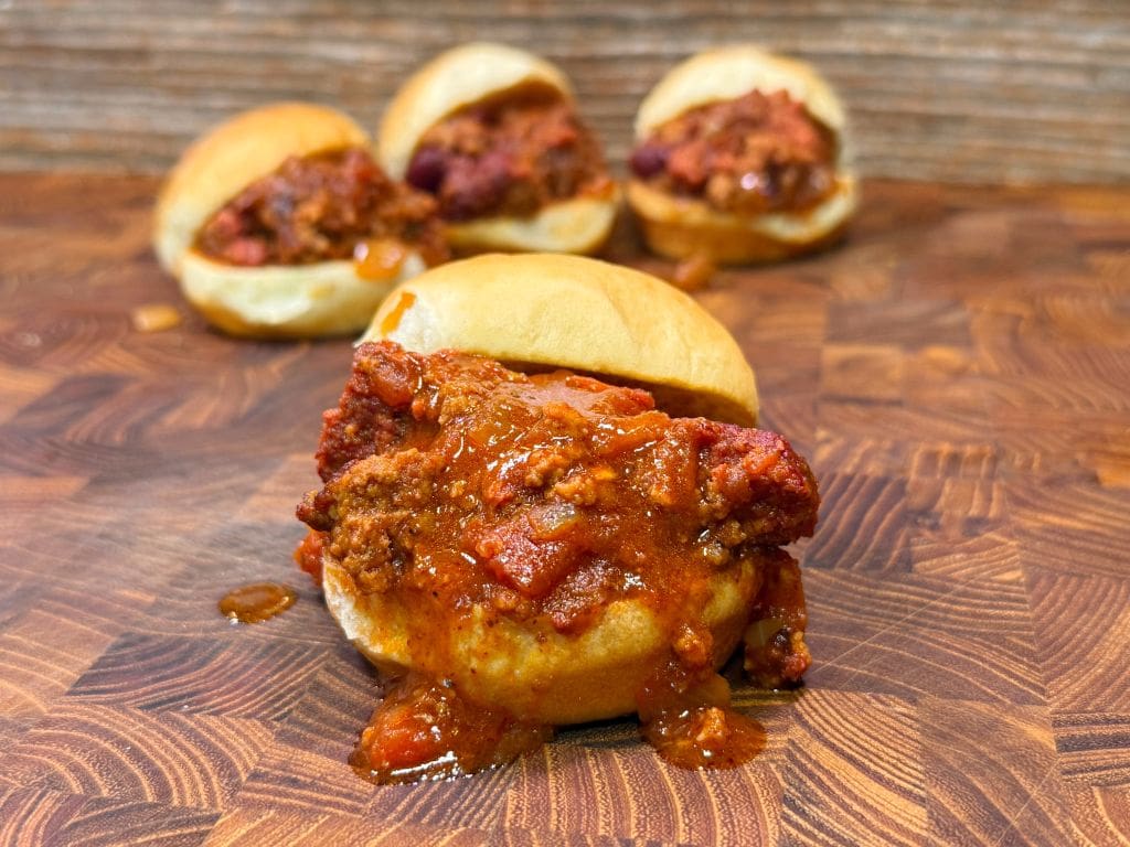 A close-up of a slider with spicy meat and sauce on a wooden surface, accompanied by three similar sliders in the background. The sauce drips slightly down the bun, adding to the appetizing appearance.