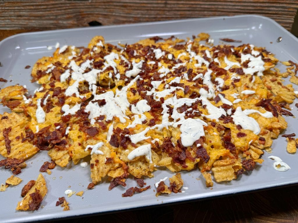 A tray of nachos topped with melted cheese, crumbled bacon, and drizzled with a white sauce, likely sour cream or ranch dressing. The tray sits on a wooden surface.