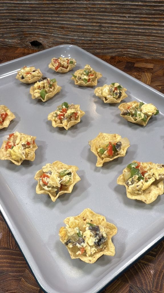 Taco chips filled with a mixture of scrambled eggs, diced red and green bell peppers, and bits of dark beans are arranged on a gray baking sheet. The background features a textured wooden surface.