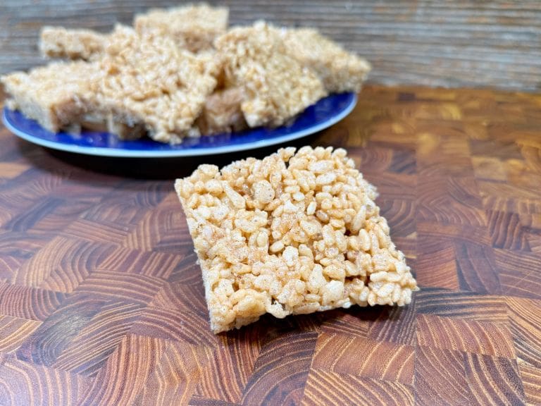 A close-up of a rice cereal treat square placed on a wooden surface, with a blue plate full of more treats blurred in the background. The treat has a golden, crispy texture.