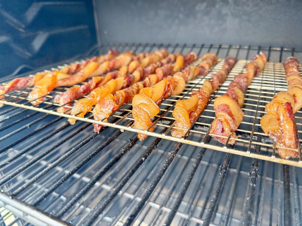 Strips of bacon wrapped around skewers are being cooked on metal racks inside an oven or smoker. The bacon is evenly spaced and appears to be caramelized. The interior of the cooking appliance is visible, showing the heating elements and walls.