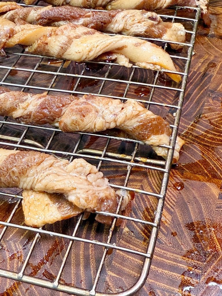 Raw marinated meat strips twisted into spirals on a wire rack over a wooden cutting board. The meat appears seasoned and is ready for cooking.