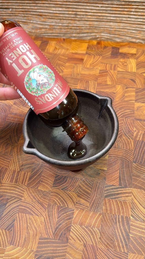 A person is pouring hot honey from a Blis hand-crafted bottle into a small, dark bowl on a wooden countertop. The countertop features a geometric pattern.