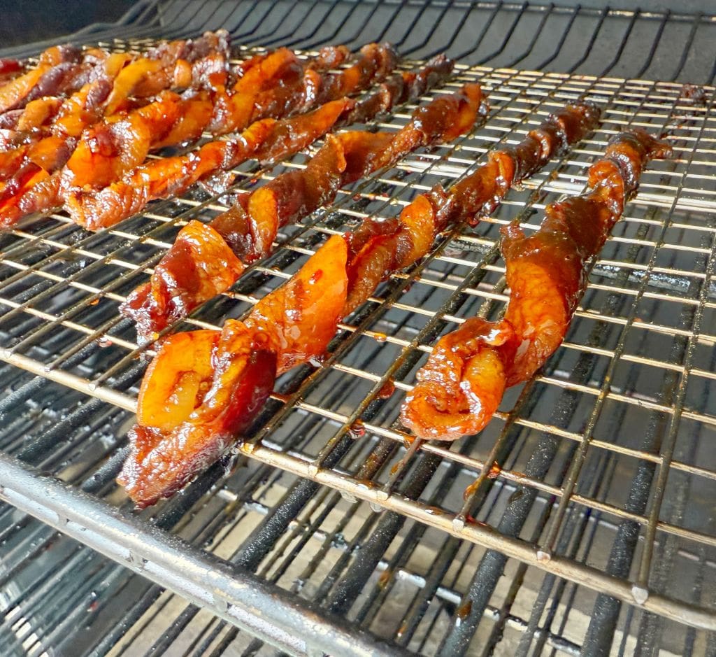 Close-up of rows of candied bacon strips on a metal rack inside an oven. The bacon appears caramelized and glazed with a dark mahogany color, giving a glossy appearance. The oven racks are visible in the background.