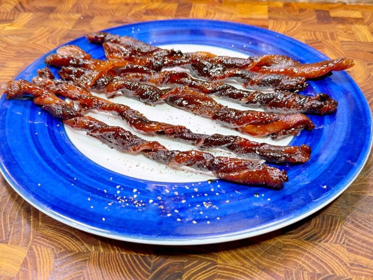 Twisted candied bacon strips arranged on a blue and white plate. The bacon is glistening with a sugary coating, placed on a wooden surface.
