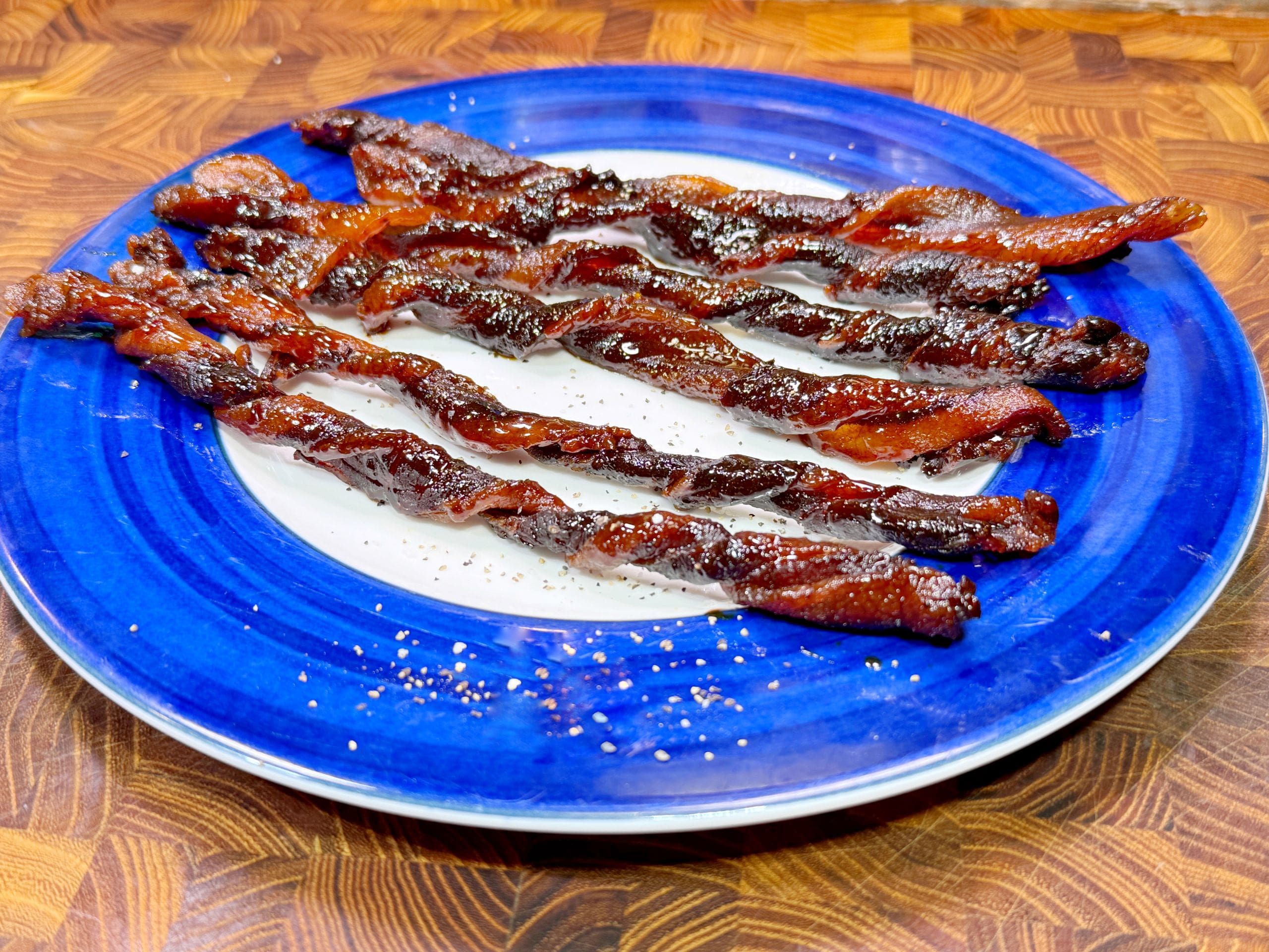 Twisted candied bacon strips arranged on a blue and white plate. The bacon is glistening with a sugary coating, placed on a wooden surface.