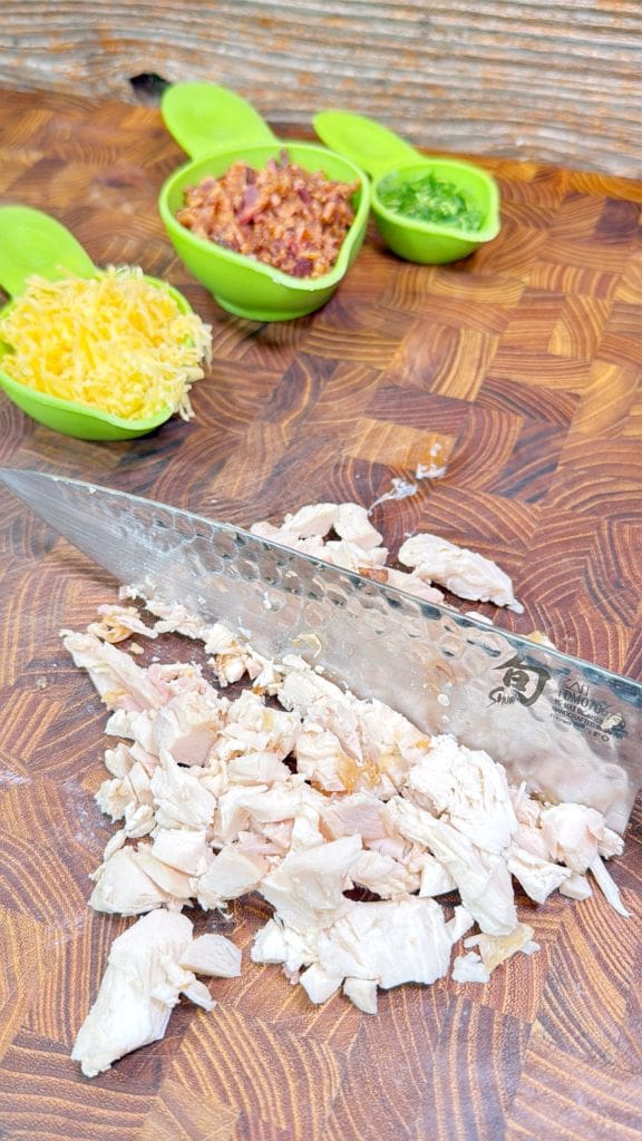 Chopped chicken on a wooden cutting board with a silver knife. In the background, three green bowls hold shredded cheese, bacon bits, and chopped parsley.