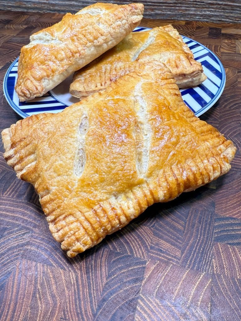 Three golden-brown puff pastry turnovers are arranged on a blue and white striped plate. One turnover is placed in the foreground on a wooden surface, showcasing its flaky texture and crimped edges.