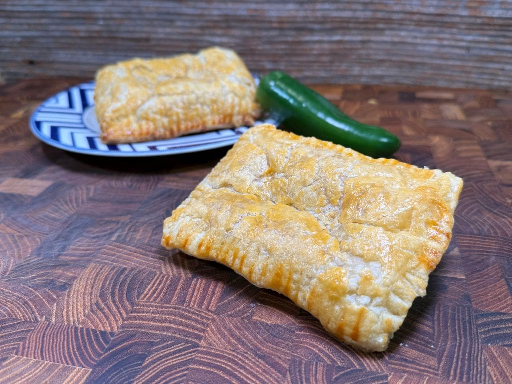 Two golden-brown puff pastry rectangles on a wooden surface, one on a blue and white patterned plate, with a green jalapeño pepper beside them. The background features a textured wooden pattern.