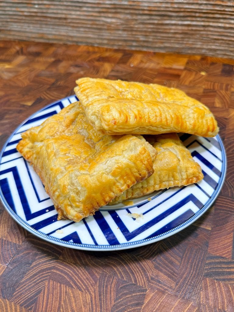 Three golden-brown puff pastries are stacked on a blue and white zigzag-patterned plate, placed on a wooden surface. The pastries have a crispy, flaky texture.