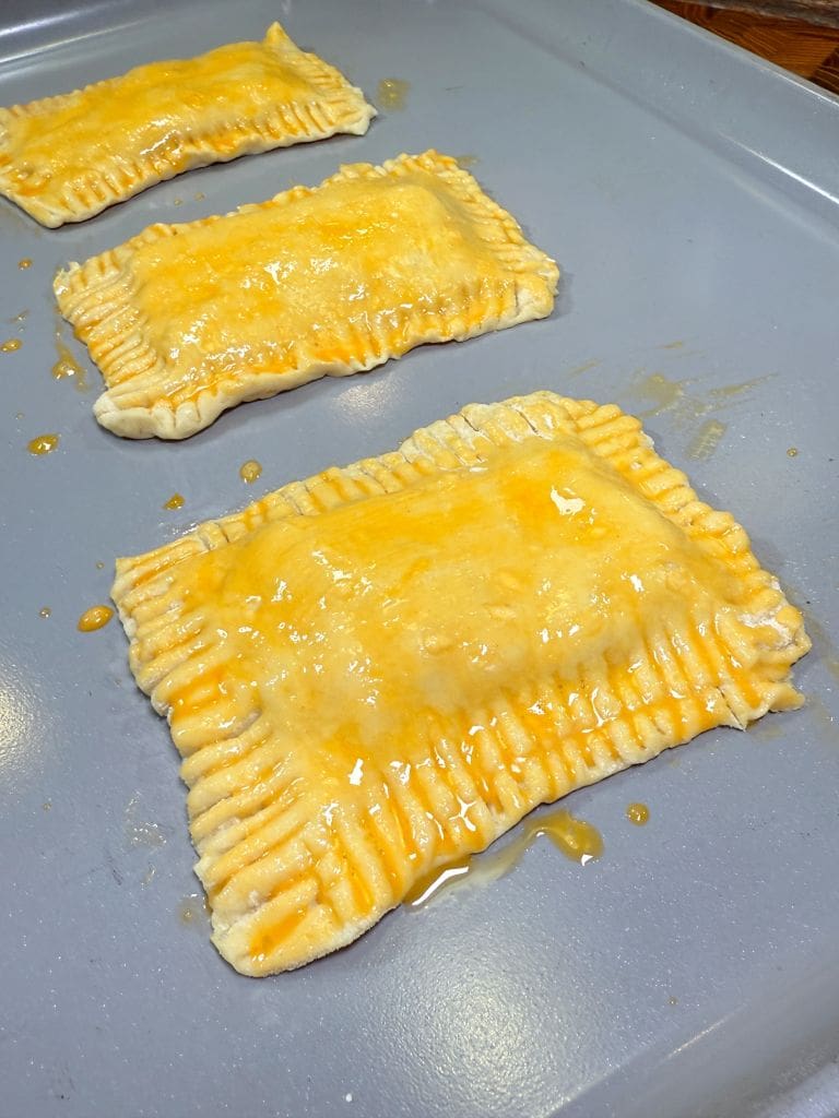 Three rectangular pastries with crimped edges and a shiny glaze sit on a baking tray. The pastries appear freshly prepared and are ready for baking.