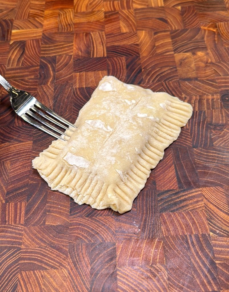 A rectangular piece of uncooked pastry with crimped edges is on a wooden cutting board. A fork is placed next to it, used for pressing the edges. The board has a rich, textured pattern.