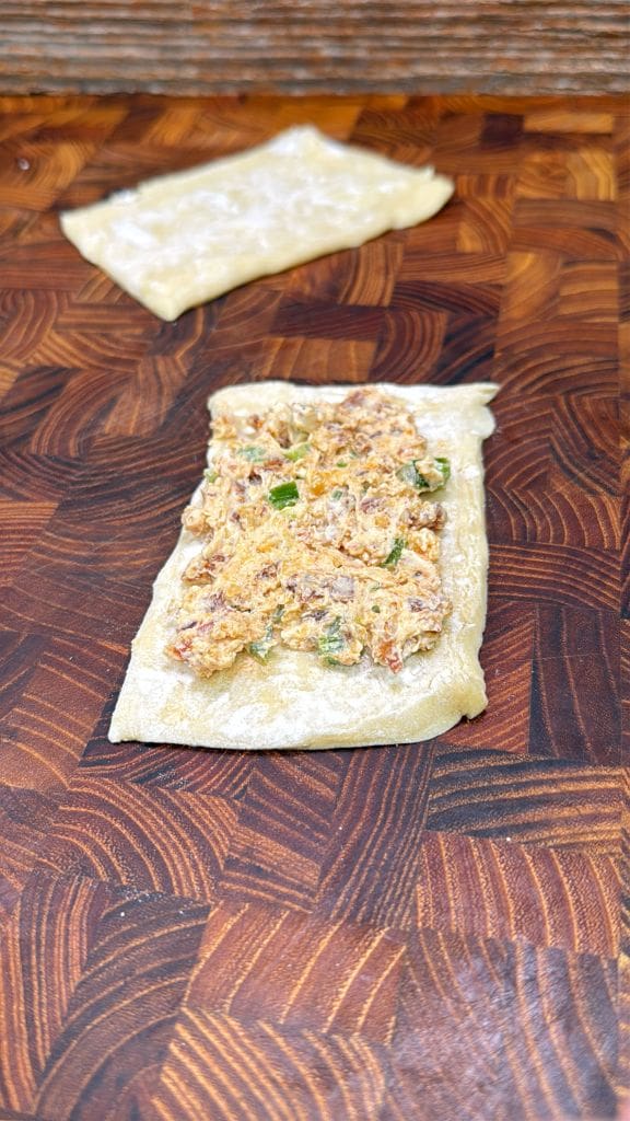 Rectangular pastry dough is topped with a savory filling on a wooden cutting board. A second piece of plain dough is in the background. The filling includes mixed ingredients like herbs and possibly cheese.