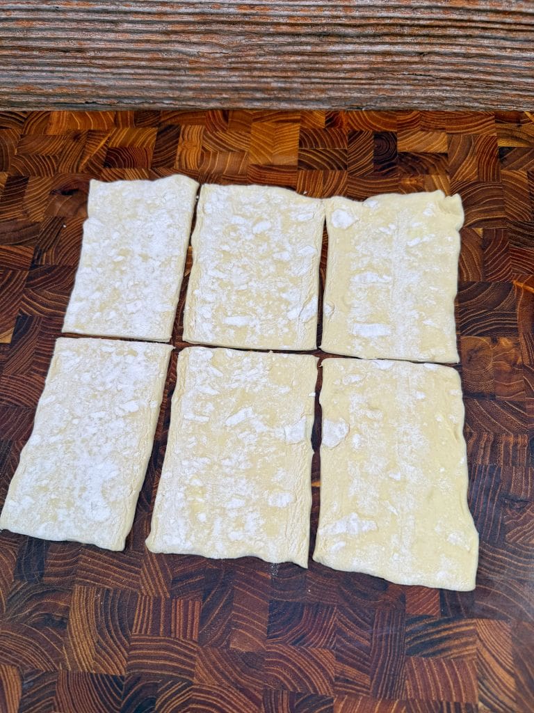Six rectangular sheets of uncooked pasta lie on a dark wooden cutting board with a textured pattern. Each sheet has a slightly uneven shape and a dusting of flour on the surface.