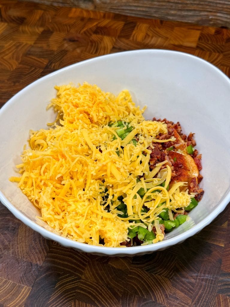 A white bowl filled with shredded cheddar cheese, diced green onions, and crumbled bacon on a wooden countertop for a jalapeno poppers recipe