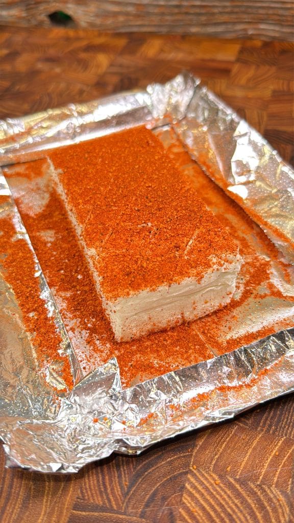 A rectangular block of cream cheese covered in red powdered seasoning, placed on a piece of aluminum foil. The foil rests on a wooden surface, and the seasoning is spread unevenly across the cheese and foil.