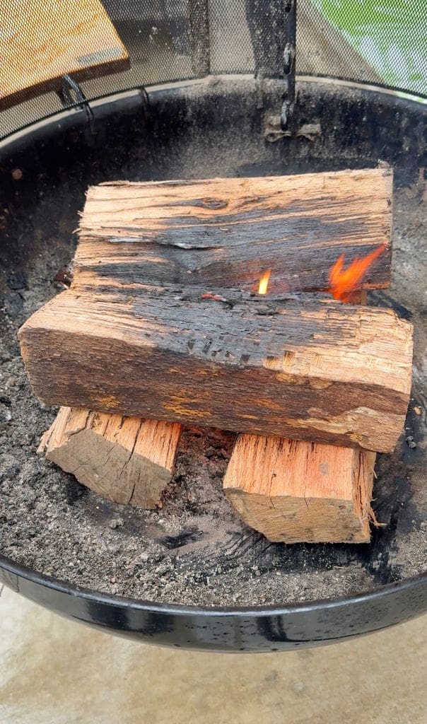 A firepit containing burning logs, with a small flame visible on top. The wood is partially charred, situated on a base of ash. The pit is surrounded by a metal screen. Ground and grass are faintly visible in the background.