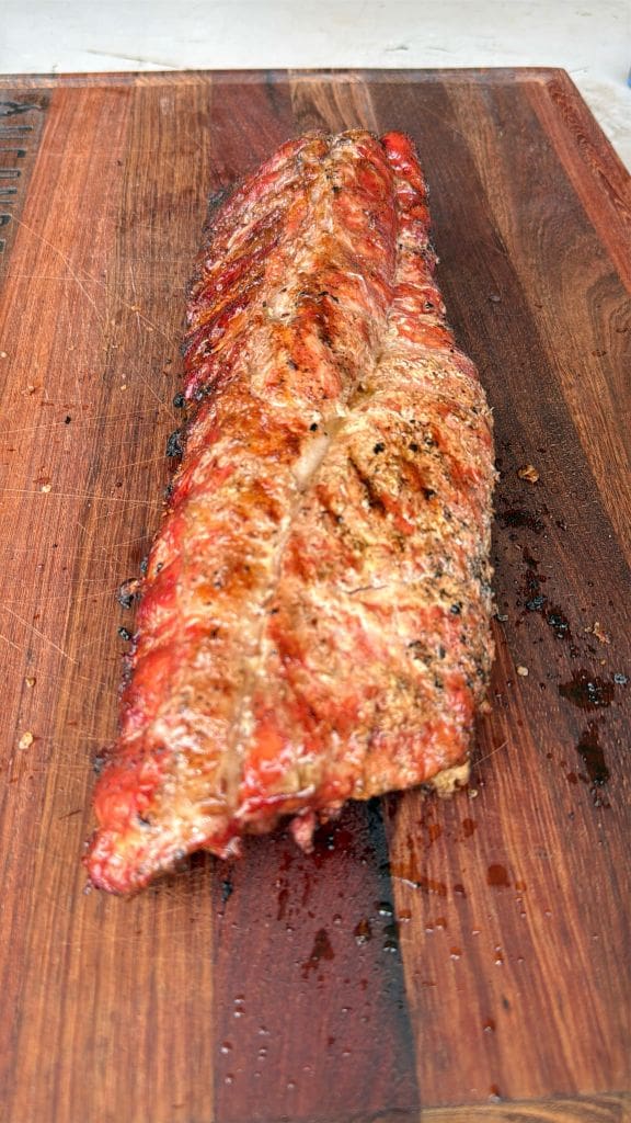 Grilled rack of ribs with a charred texture, resting on a wooden cutting board. The ribs are cooked on a campfire and are glazed and appear juicy, with visible grill marks and lightly seasoned with herbs and spices.