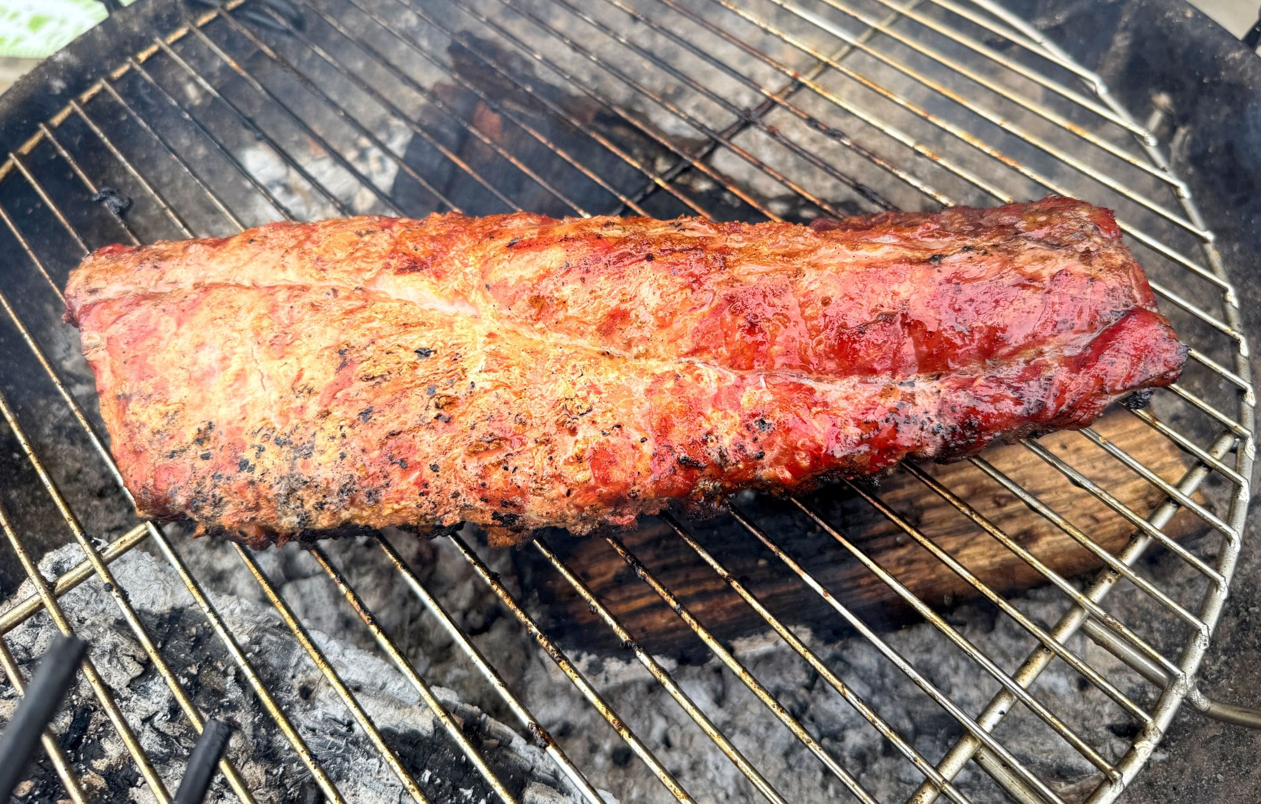 A slab of seasoned pork is cooking on a round grill over hot coals. The meat is charred and glistening, suggesting it is being smoked or barbecued. The grill grates and pieces of wood are visible underneath the pork.