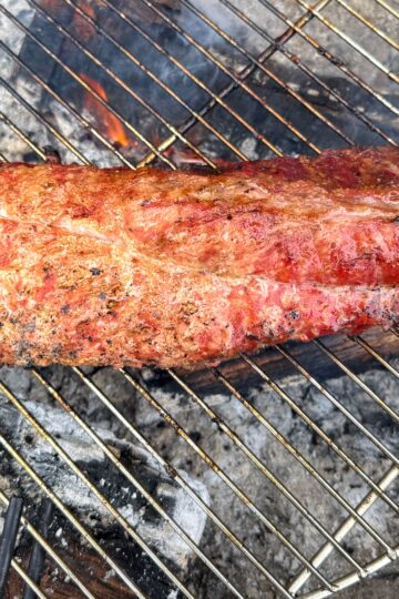 A piece of seasoned steak is grilling over hot charcoal on a round barbecue grill, with flames and smoke rising from beneath. The meat is cooked to a reddish-brown, showing grill marks.