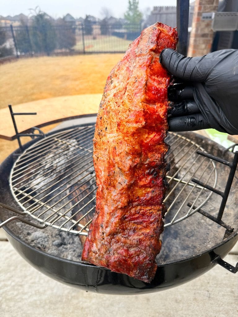 A hand wearing a black glove holds a rack of grilled ribs over a round barbecue grill. The ribs are cooked to a golden-brown, suggesting they are smoked. In the background, there is a yard with a fence and slightly blurred surroundings.