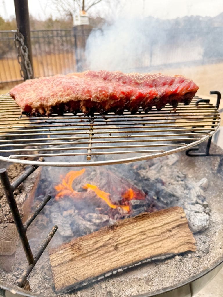 Ribs coated with sauce are grilling on a round grate over an open flame. Smoke rises from the fire, and a piece of wood is burning below, creating a rustic cooking scene. A metal fence and overcast sky are visible in the blurred background.