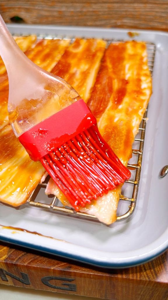 A close-up of bacon slices on a wire rack over a tray. A red silicone brush is being used to apply a shiny glaze over the bacon. The glaze has a rich, glossy appearance.