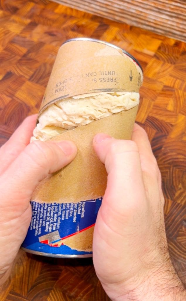 Two hands are opening a cardboard tube of crescent rolls on a wooden surface. The packaging is partly peeled, revealing the dough inside. The persons thumbs are applying pressure along the seam to open it.