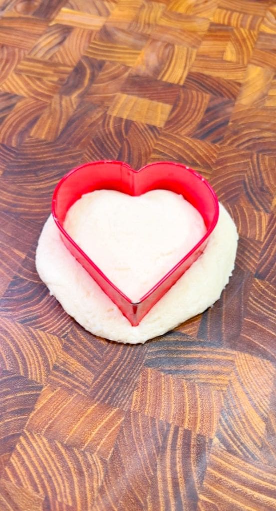 A red heart-shaped cookie cutter sits on a piece of dough placed on a wooden countertop with a checkered pattern.