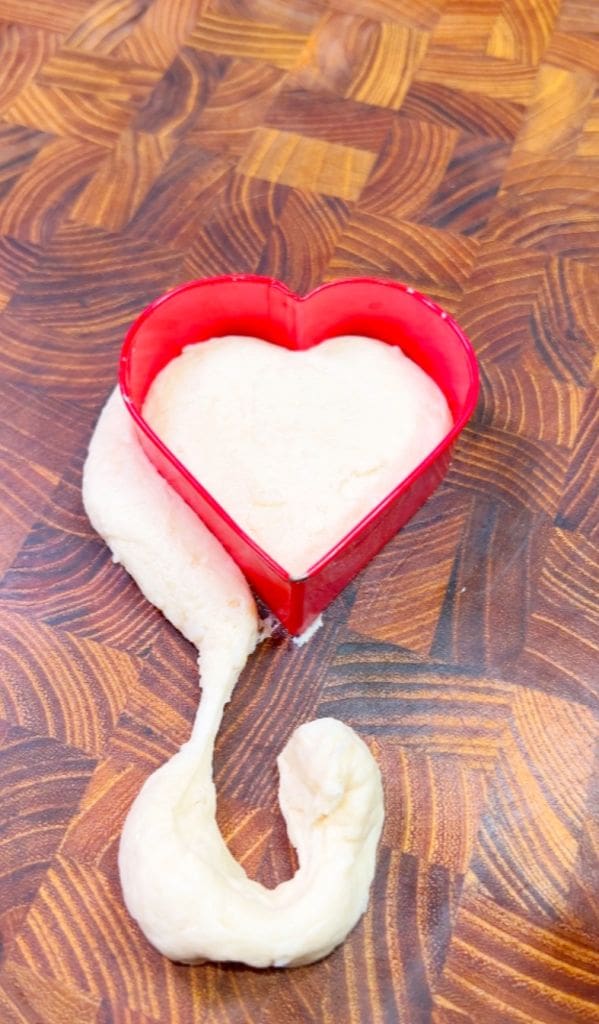 A heart-shaped red cookie cutter filled with white dough sits on a textured wooden surface. Some dough spills out from the bottom, creating a tail-like shape.
