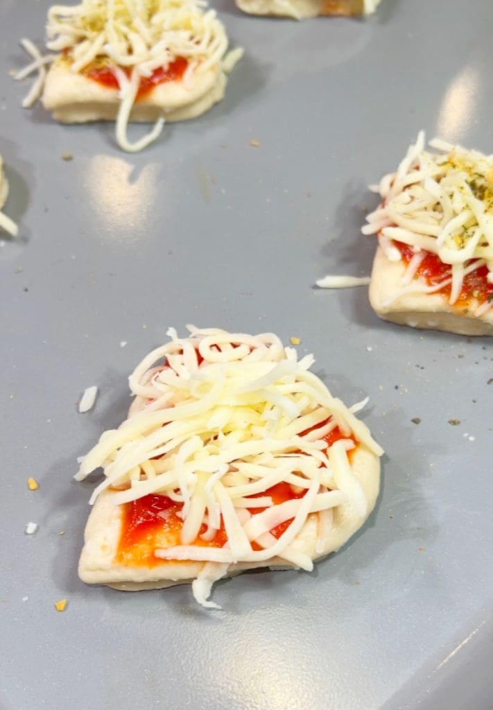 Small dough pieces topped with red sauce and shredded cheese are arranged on a gray tray, ready for baking. Some of the cheese spills over the sides of the dough.