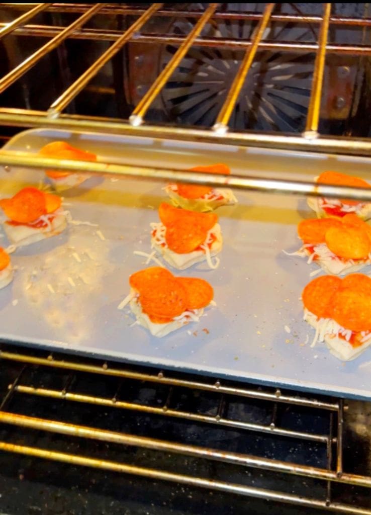 Mini pizzas with shredded cheese and pepperoni are baking on a silver tray inside an oven.