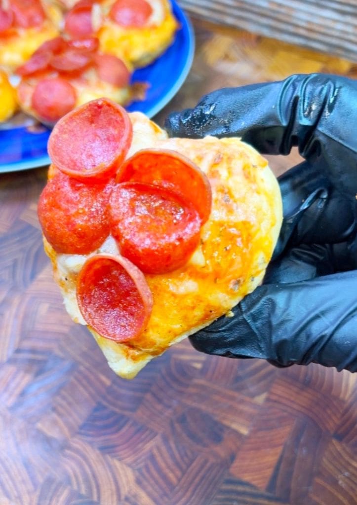 A hand in a black glove holds a heart-shaped mini pizza topped with pepperoni slices. A plate with more mini pizzas is visible in the background on a wooden surface.