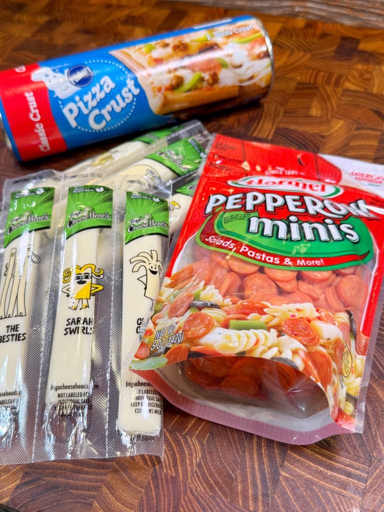 A wooden table displays pizza-making ingredients: a can of pizza crust dough, a package of mini pepperoni slices, and three packs of string cheese with playful packaging designs.