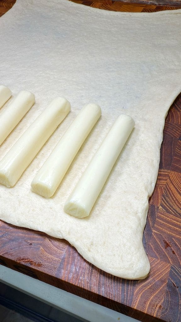 Rolled-out dough on a wooden surface with five long sticks of cheese placed on it, ready for wrapping.