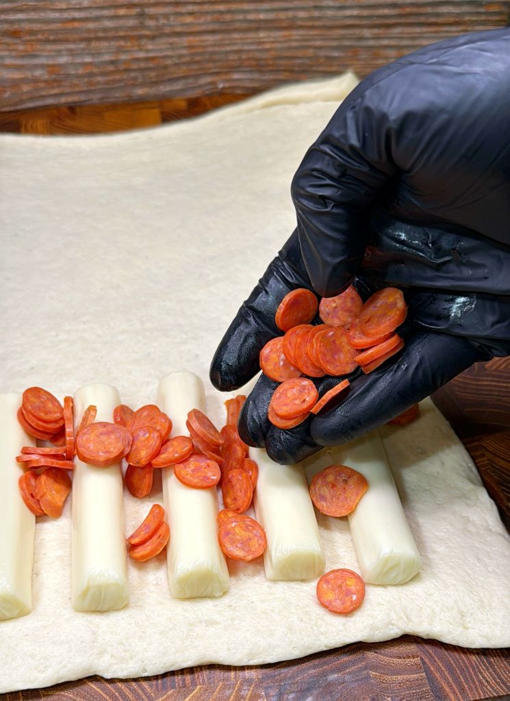 A person wearing black gloves places slices of pepperoni over string cheese sticks arranged on a dough sheet. The scene suggests preparation for a snack or appetizer, using fresh ingredients on a wooden surface.