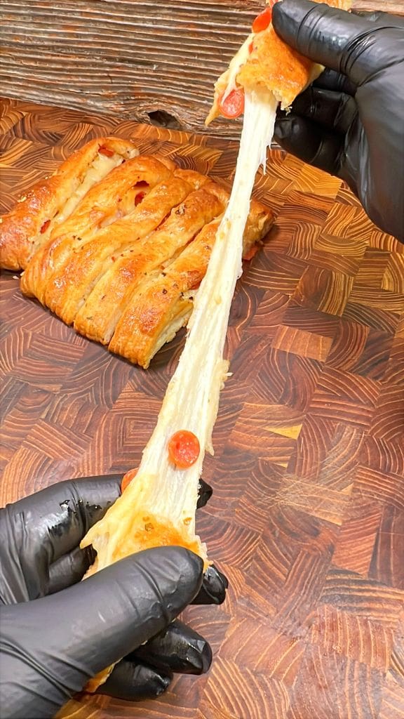 Close-up of hands tearing apart a cheesy pepperoni pizza bread braid on a wooden cutting board. The cheese is stretching between the two pieces, and the person is wearing black gloves.