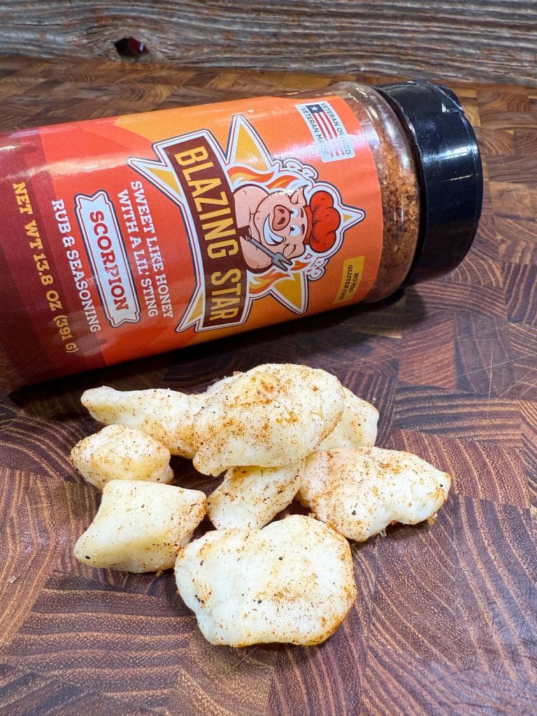 A container of Blazing Star seasoning labeled Scorpion lies on its side, spilling some seasoning powder onto puffed pork skins on a wooden surface. The pork skins are dusted with the seasoning.