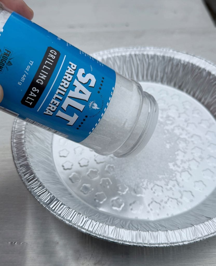 A hand is pouring salt from a container labeled Grilling Salt into a round, disposable aluminum pie pan. The pan is on a white surface. The salt granules are visible against the shiny interior of the pan.