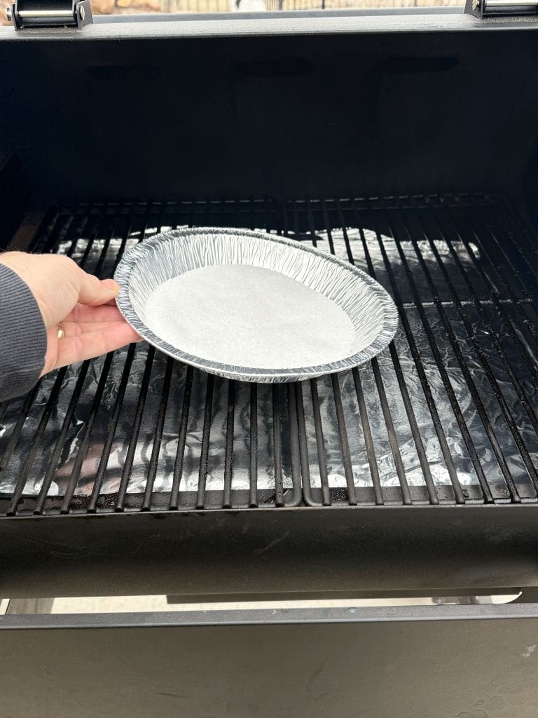A hand is placing an empty, round aluminum foil pan on the grate of a black grill. The grill appears to have foil covering the grates underneath the pan.  Getting ready to smoke salt