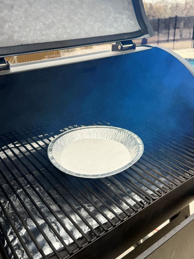 An empty foil pie pan sits on the grill grates inside a black outdoor smoker. Light smoke is visible, and the grill lid is partially open. The background shows a blurred outdoor setting.