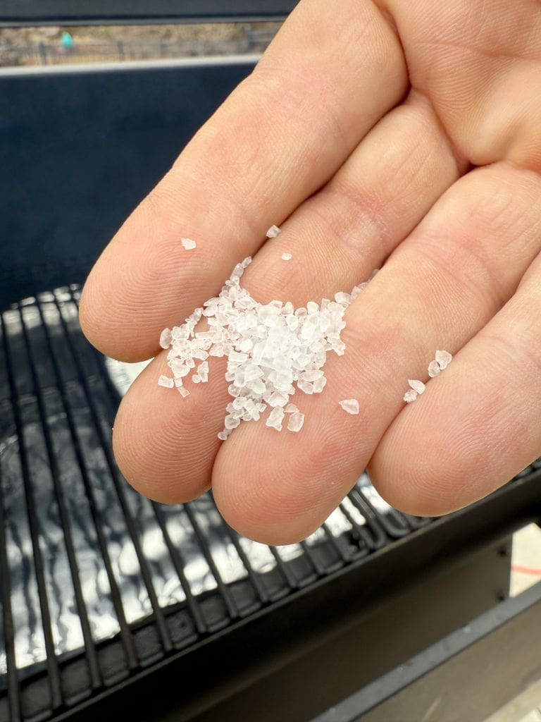 A close-up of a hand holding coarse sea salt crystals over a grill. The grill grates are visible in the background, suggesting preparation for cooking or seasoning food.