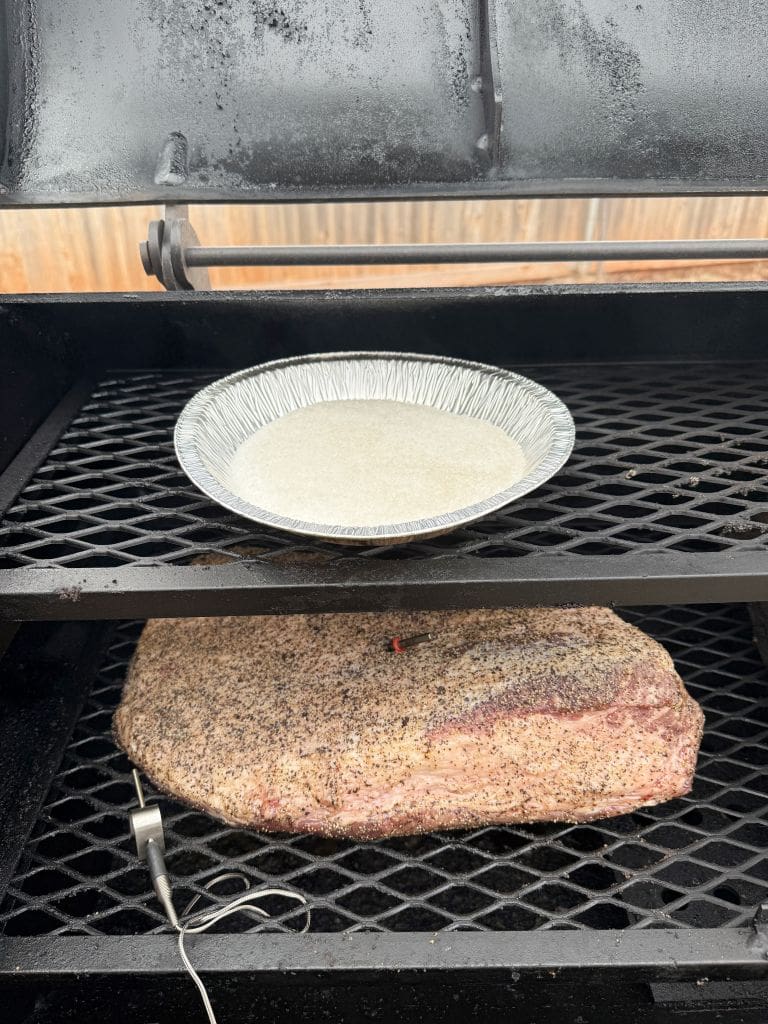 A meat brisket seasoned with pepper is being smoked on a grill. A metal probe for a thermometer is inserted into the brisket. Above, a foil pan with liquid sits on a grate. The grills lid is open, revealing the setup.