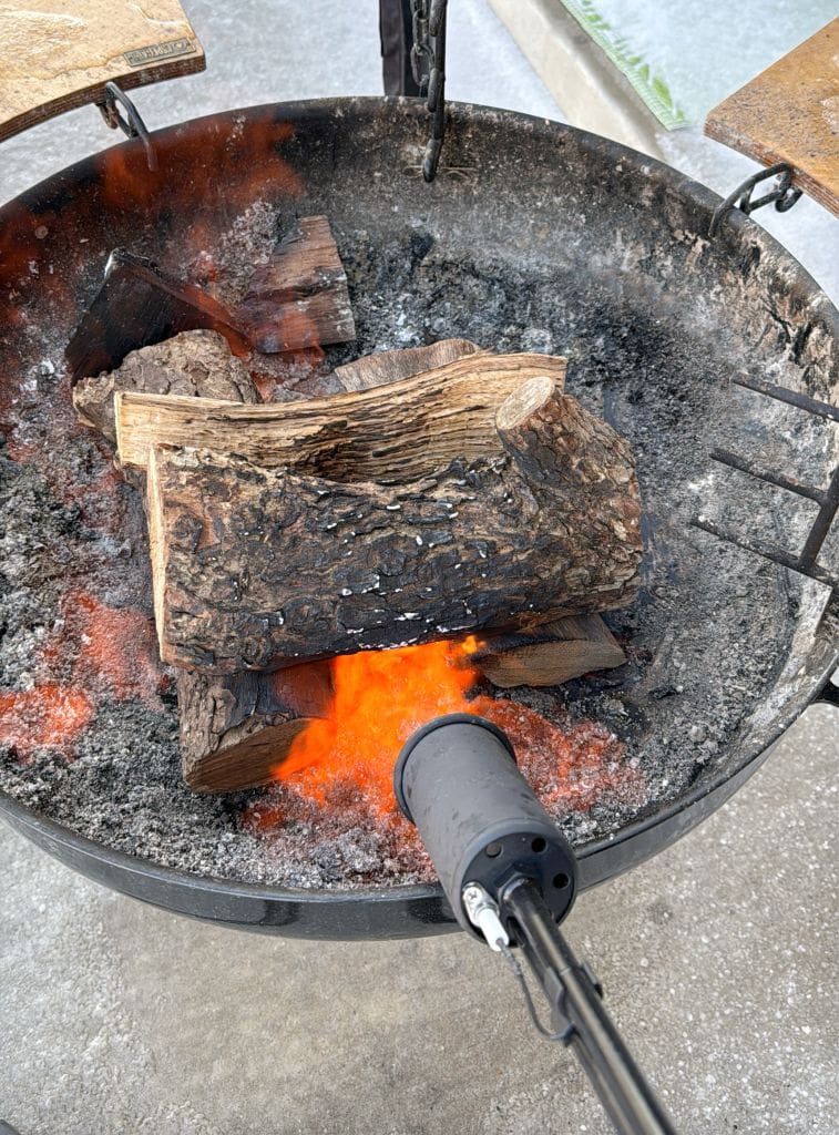 A fire pit filled with ash and charred wood is being ignited by a flame torch. Bright orange flames are visible under the stacked logs, while metal supports surround the pit on a concrete surface.